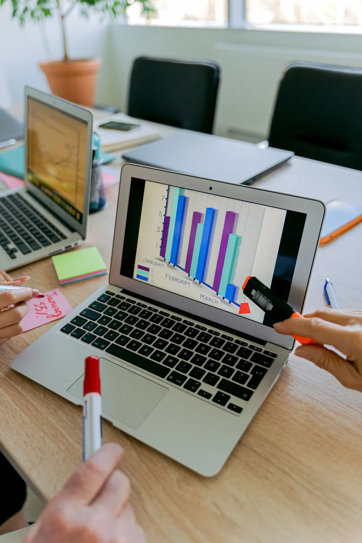Laptop screen showing a bar chart, with hands holding a marker and a highlighter at a shared desk