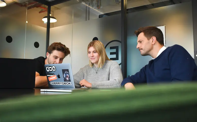 Three colleagues gathered around an open laptop in a bright meeting room, looking at the screen together
