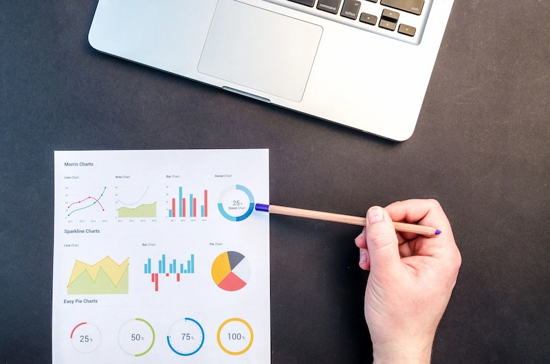 Printed charts and graphs on paper beside a laptop, with a hand holding a pencil